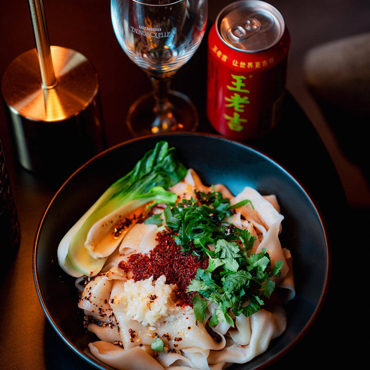 Schüssel mit Nudeln, Gemüse und Gewürzen; Glas und Getränkedose daneben; warme Beleuchtung.