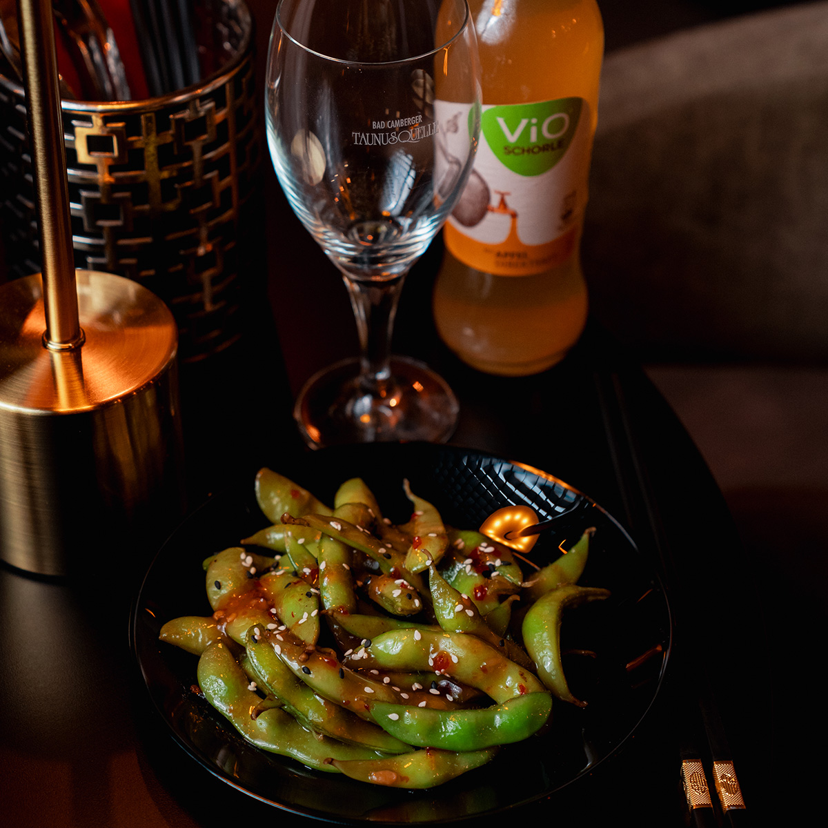 Schwarzer Teller mit gewürzten grünen Edamame, daneben Glas und gelbes Getränk. Stimmungsvolle Beleuchtung.