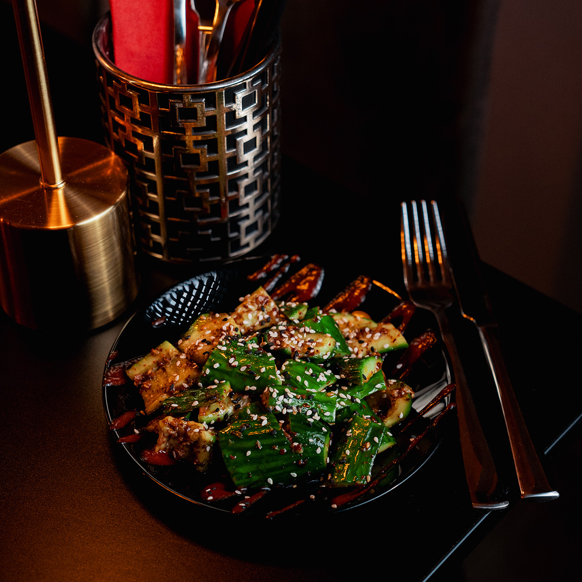 Ein schwarzer Teller mit gegrilltem Gemüse, bestreut mit Sesam, daneben Besteck auf schwarzem Tisch.