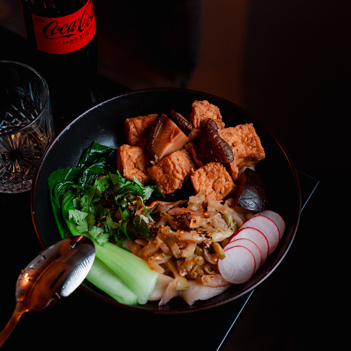 Eine Schüssel mit Tofu, Shiitake, Gemüse und Radieschen; daneben eine Cola-Flasche.
