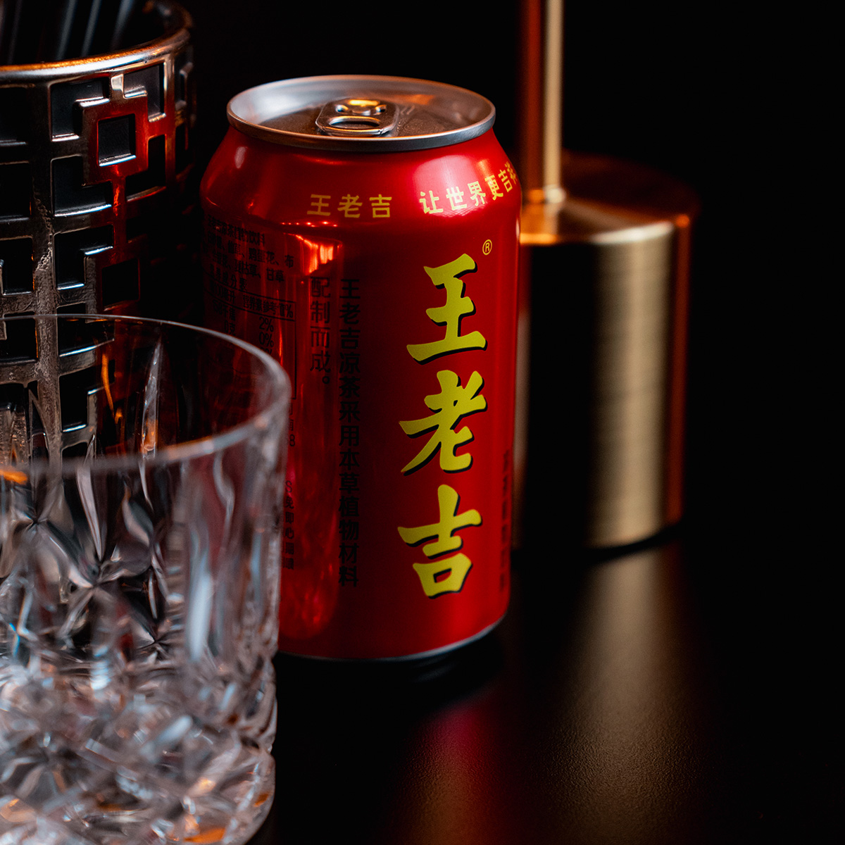 Rote Getränkedose mit asiatischer Schrift neben Kristallglas und schwarzem, gemustertem Behälter.