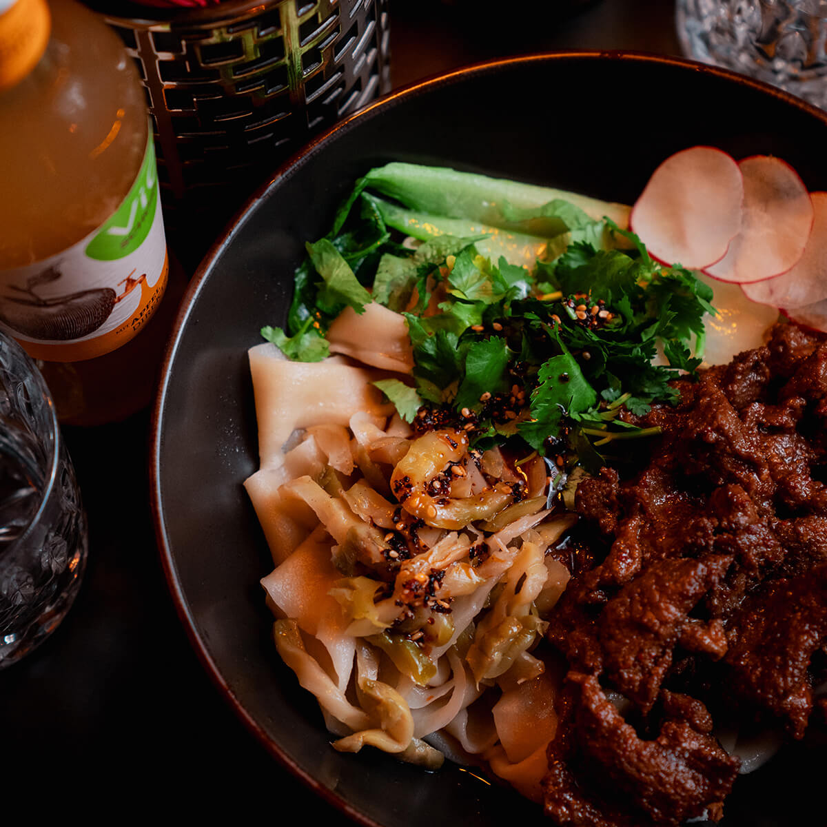 Schüssel mit Nudeln, Fleisch, Gemüse und Gewürzen, daneben eine Flasche Saft; lebendige Farben.